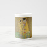 Älskare Kissing Embrace av Gustav Klimt Benporslin Mugg<br><div class="desc">Solljus blundar från det virvlande guldomfamning som omger älskare i Gustav Klimts mästerverk, The Kiss. Scenen är en paradox med intimitet och isolering. Paret står på en avgrund, en fält av färgstarka blommor på fötterna, men ändå är de omedvetna mot omvärlden. Deras blickar är låsta, deras kroppar är sammanflätade i...</div>