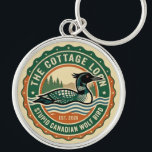 Cottage Loon Stupid Canadian Wolf Bird  Rund Silverfärgad Nyckelring<br><div class="desc">Circular badge with a central illustration of a waterfowl floating on stylized blue and cream waves. The bird features a dark head, a pointed beak, and a distinct white checkered pattern on its back. The background consists of green pine tree silhouettes against a pale sky. The design is framed by...</div>