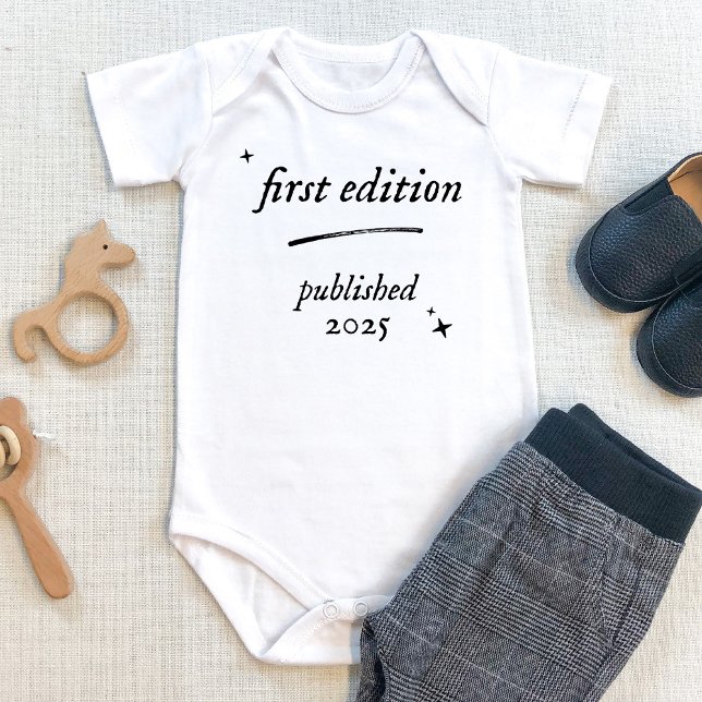 Första utgåvan, Bok, utgiven 2025 Baby-kroppsdräkt T Shirt (First edition baby bodysuit with personalized birth year. Shown on beige table with baby pants. book)