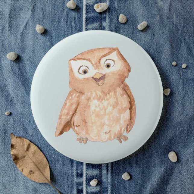 Glad Uggle Akvarell Knapp (A watercolor owl cartoon smiling on a pale blue pin, laid on denim amid pebbles and leaves.)