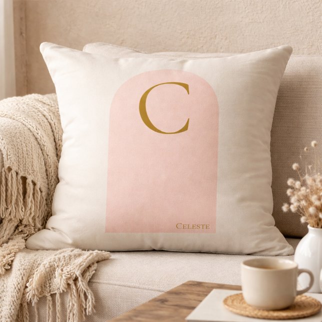 Kastkudde med monogram och rosafärgad valv kudde (Soft ivory meets blush arch detail, finished with a gold serif monogram for refined neutral living.)