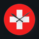 Schweiz flagga rund klocka<br><div class="desc">Schweiz flagga (tysk: Schweizerfahne; Fransk: drapeau de la Suisse; italiensk: bandiera svizzera; Romansh: bandiera de la Svizra) visningar ett vitt kor mitt i ett kvadrat rött fält. Det vita kor är känt som det schweiziska kor.</div>