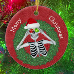 Skeleton i Gräs, frisk, rödhatt Julgransprydnad Keramik<br><div class="desc">Det här hängda keramiska prydnadsämnet har ett lustigt skelett som håller prydnadsföremål med snöflingor fram till ögon och bär en röd julhatt inuti en grönt-nålkrans på djup röd bakgrund. Anpassa texten på baksidan med hjälp av mallarna och God jul på framsidan.</div>
