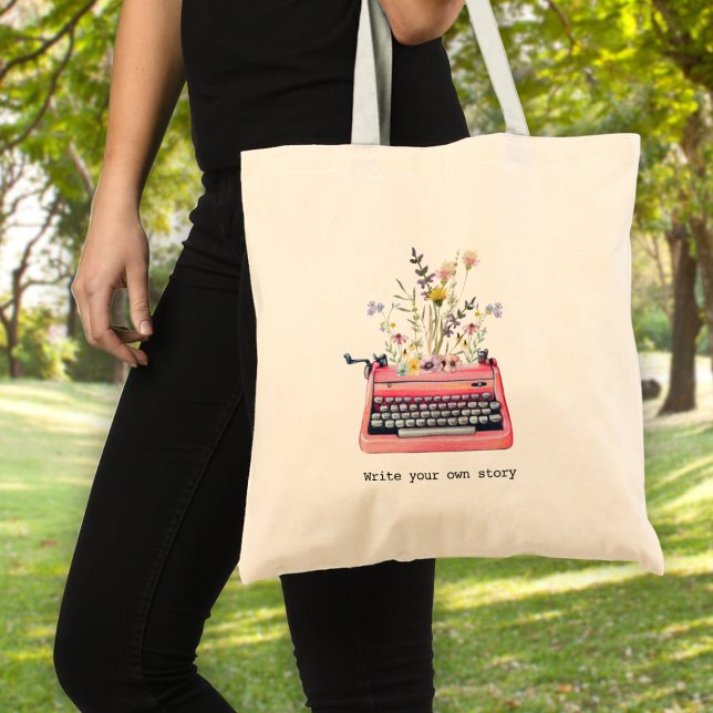 Skriva din egen artikel, skrivprogram tygkasse (A woman holding a tote bag with a typewriter graphic and wildflowers & text: "write your own story")