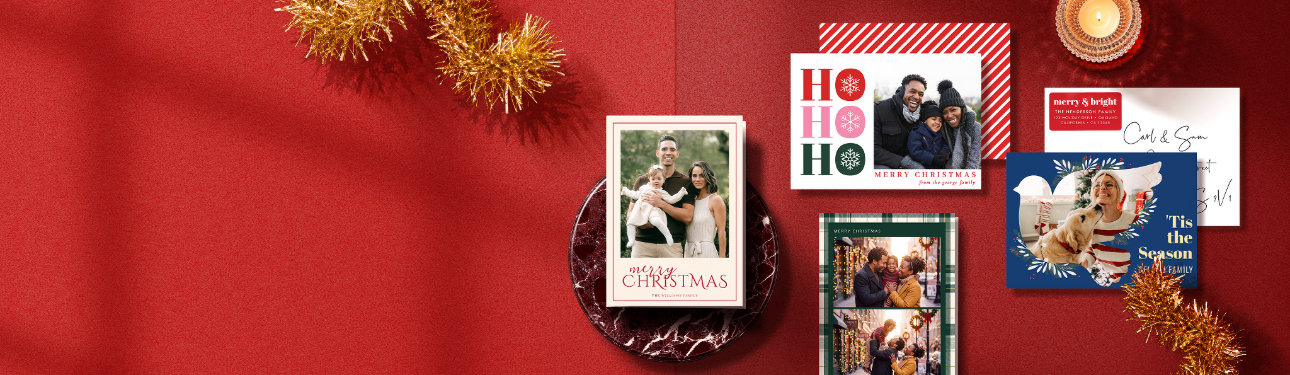 Personalised Christmas photo cards in gold, green, and navy designs arranged on a festive red background with tinsel and candlelight.