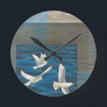 Tre vita Fiskmåsar som flyger över Vatten Rund Klocka<br><div class="desc">Behöver du komma undan? Gör det genom att välja den här målningen av konstnären Tara Friel som representerar tre fiskmåsar som flyger över vatten. Synen på havets vågar och det levande färger gör denna utformning mycket realistisk.</div>