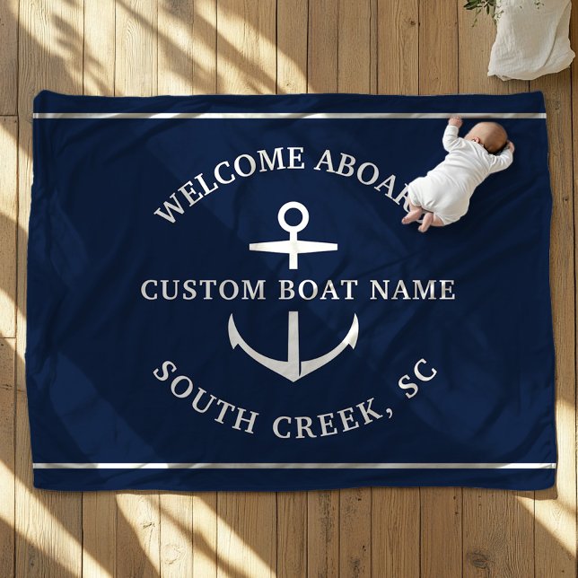 Välkomstankare för den moderna Anpassningsbarna Bo Fleecefilt (A baby, dressed in white, lies on a navy blue blanket with an anchor motif. Text is Welcome Aboard")
